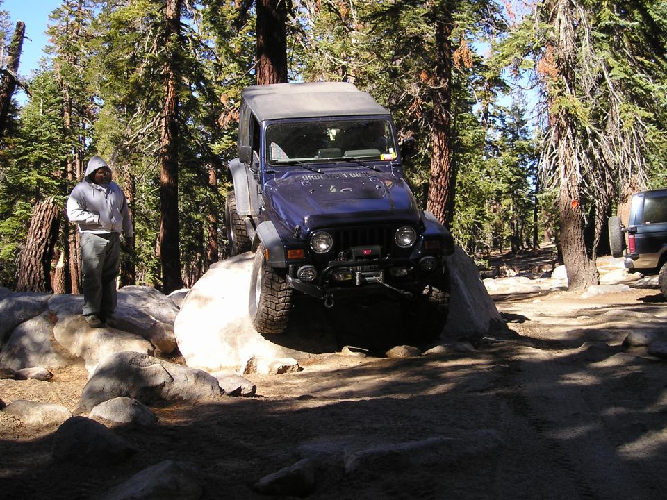 Dark blue Jeep Wrangler TJ navigating rocks on a wooded trail. Person stands on rocks nearby