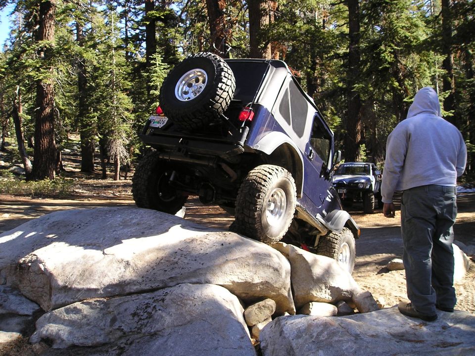 Blue Jeep Wrangler navigating large rocks on a trail, another Jeep visible behind