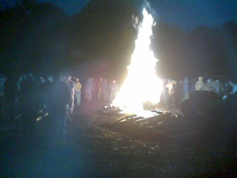 1976 GMC C/K Pickup - A large bonfire illuminates a crowd of people gathered outdoors at dusk.