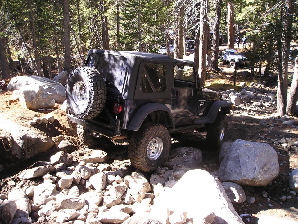 Black Jeep Wrangler with soft top and spare tire on rear, navigating a rocky forest trail