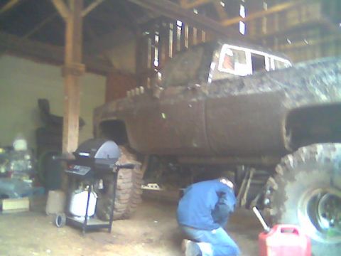 1976 GMC C/K Pickup - A person works on a heavily mud-covered pickup truck inside a barn, with a grill visible nearby.