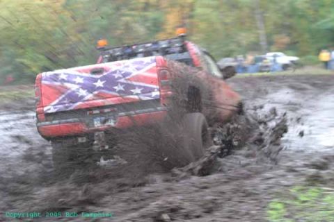 1976 GMC C/K Pickup - A red pickup truck with a Confederate flag on its tailgate is shown driving through a muddy area, kicking up a lot of mud.
