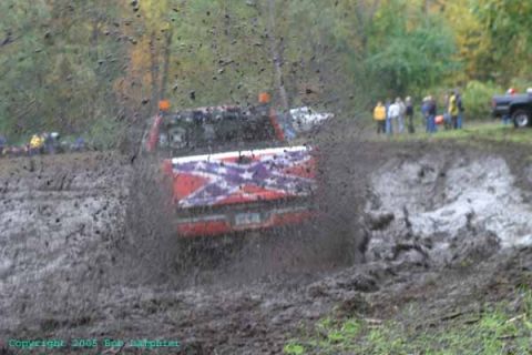1976 GMC C/K Pickup - A red pickup truck with a confederate flag decal navigates a muddy track, kicking up a large spray.