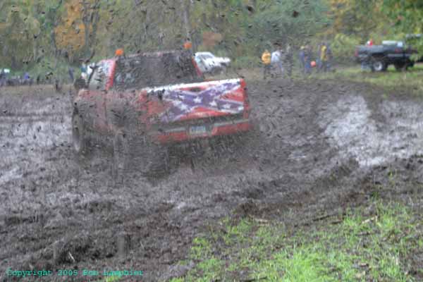 Red pickup truck with Confederate flag decal on tailgate driving through mud, other vehicles and people in background