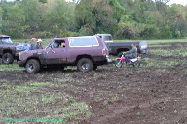 Purple SUV and a black pickup truck in a muddy field. A guy rides a dirt bike next to the SUV. Some people stand around the truck