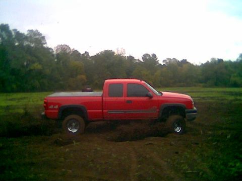 1976 GMC C/K Pickup - A red pickup truck is shown in a muddy field with trees in the distance under an overcast sky.