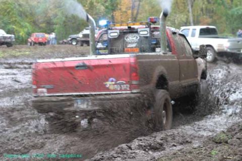 1976 GMC C/K Pickup - A red pickup truck navigates a muddy track, kicking up spray from its tires.