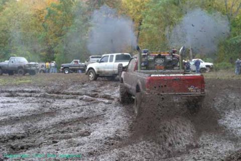 1976 GMC C/K Pickup - A red pickup truck churns up mud as it drives through a muddy field, with other vehicles and spectators visible in the background.