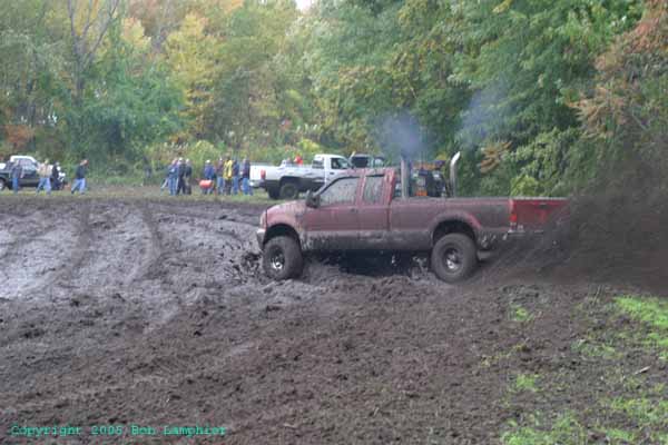 Red pickup truck with large exhaust stacks driving through mud, kicking up dirt. Other trucks and people are visible in the background