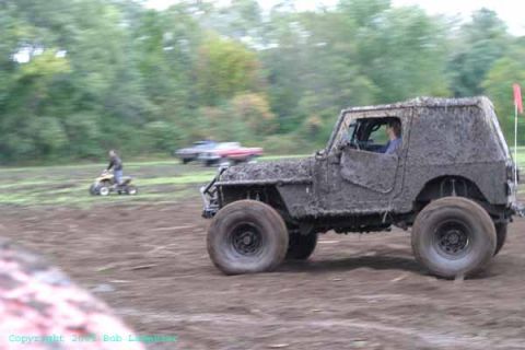 1976 GMC C/K Pickup - An off-road vehicle covered in mud navigates a muddy terrain, with other vehicles visible in the distance.