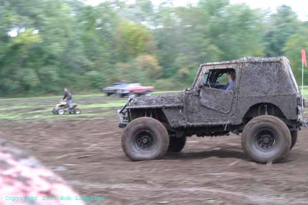 Muddy off-road vehicle driving through a field with an ATV and another vehicle in the background