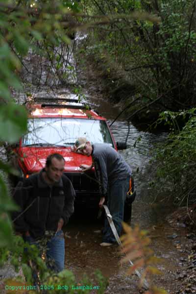 1976 GMC C/K Pickup - Two men attempt to free a red Jeep Cherokee stuck in a muddy stream.