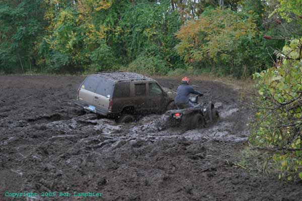 Gold SUV stuck in deep mud with an ATV rider nearby in a churned field