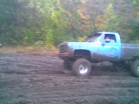 1976 GMC C/K Pickup - A blue pickup truck is driving through a muddy area with trees in the background.
