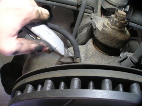 2000 Ford Excursion - Close-up of a brake rotor and suspension components with a hand holding a utility knife near a brake line.