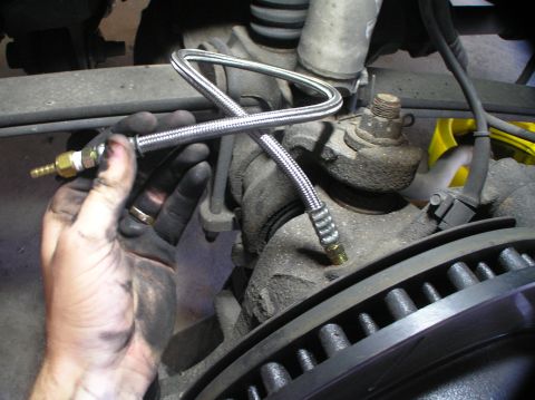 2000 Ford Excursion - A braided metal brake line is held by a dirty hand, positioned near the brake rotor and suspension of a vehicle.