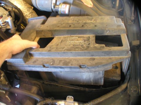 2000 Ford Excursion - A hand lifts a black plastic battery tray cover with warning labels.