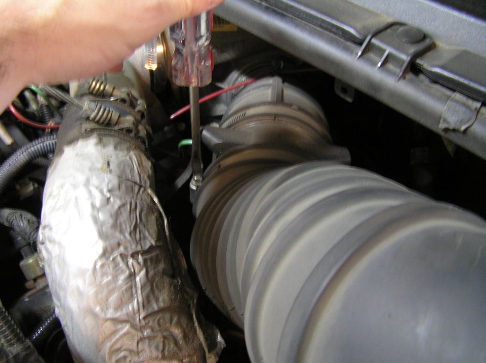 Engine bay with gray intake hose, foil-wrapped pipe, and hand holding a screwdriver