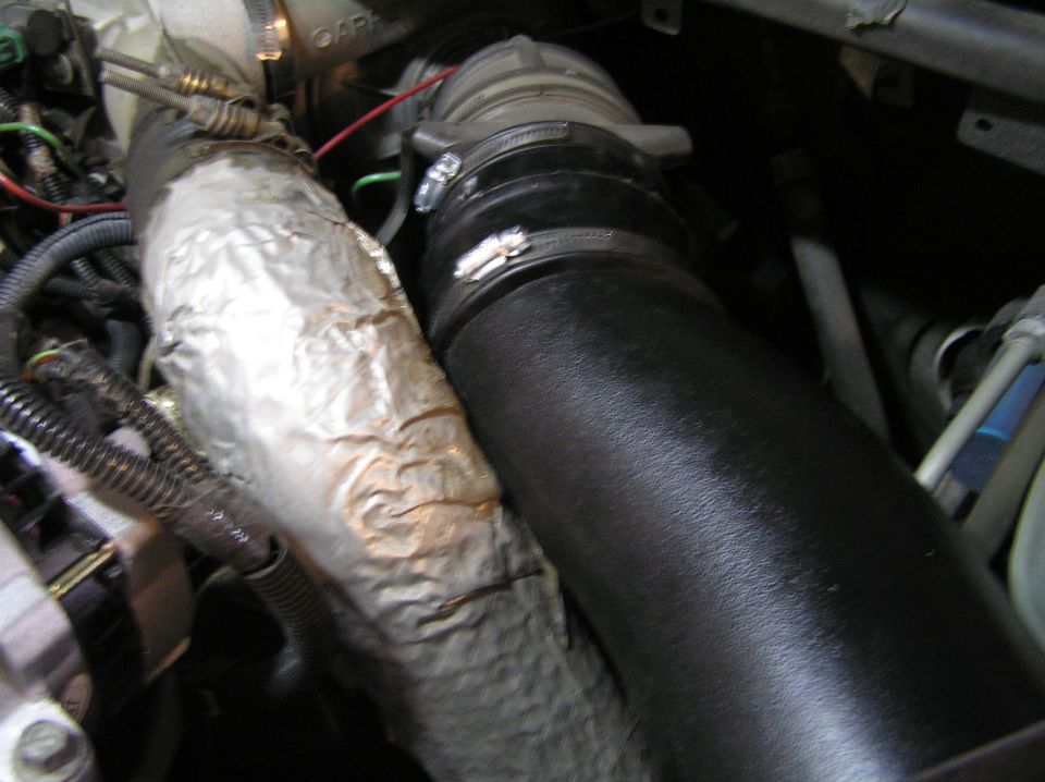 Black rubber intake hose with band clamps beside a foil heat-wrapped pipe in an engine bay