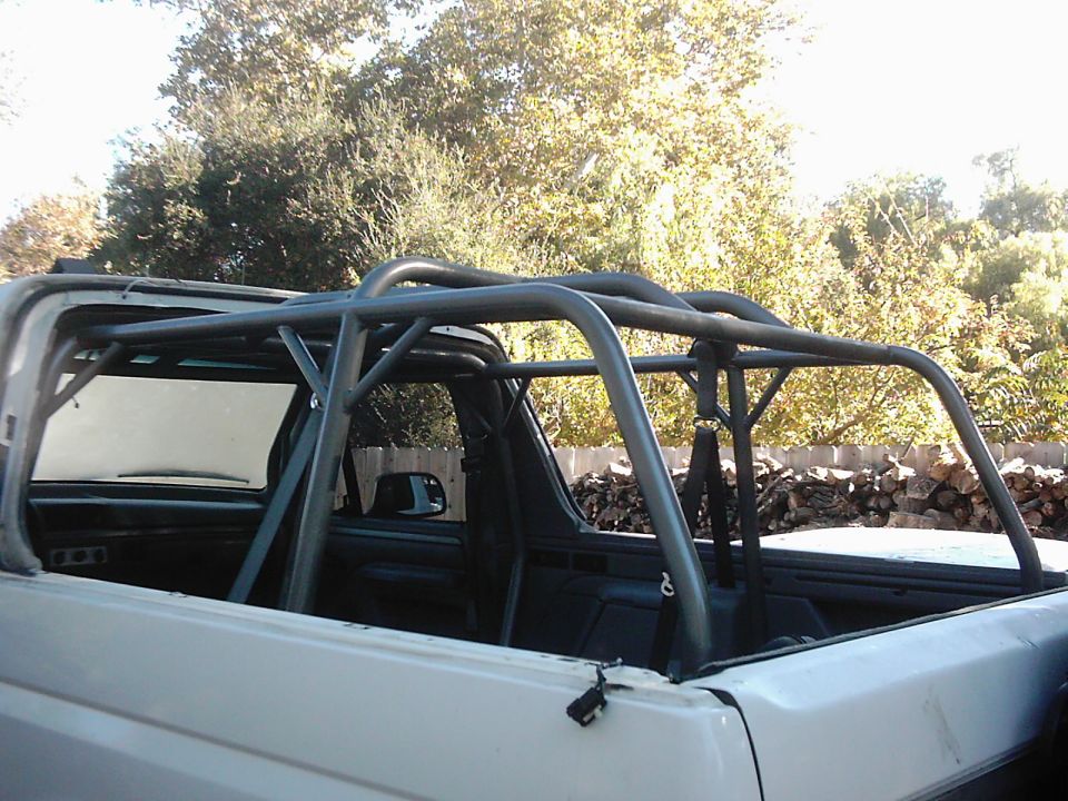 Interior view of a white Ford Bronco with a custom roll cage installed. The rear of the vehicle is visible, showing the bed and a pile of firewood in the background