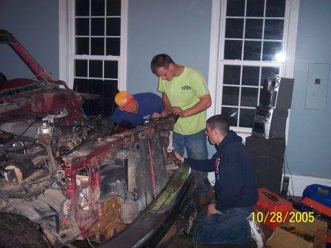 1994 Ford Bronco - Three men examine the front end of a heavily damaged red vehicle, possibly during a teardown or repair process.