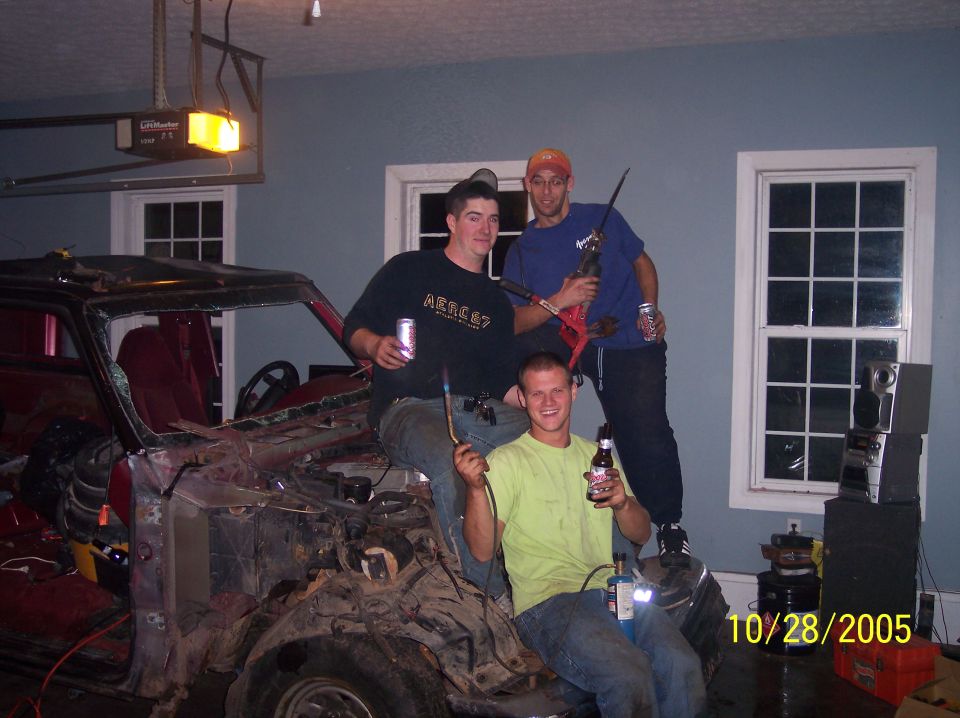 Three men are gathered around a heavily dismantled vehicle, one holding a rifle, another a welding torch. The date 10/28/2005 is visible