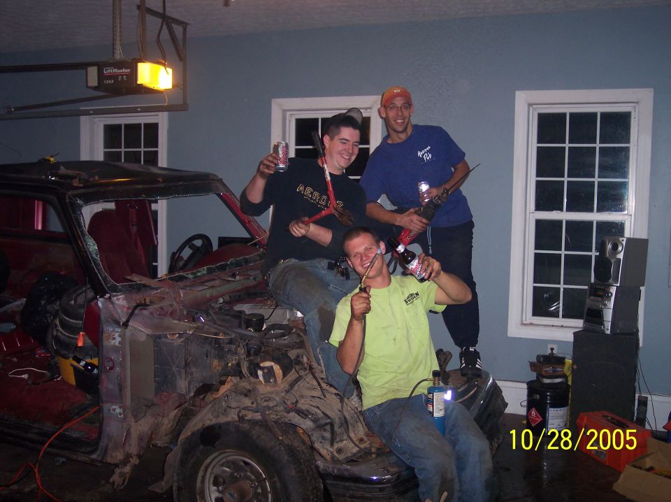 Three guys are gathered around a stripped-down vehicle in a garage, holding tools and drinks