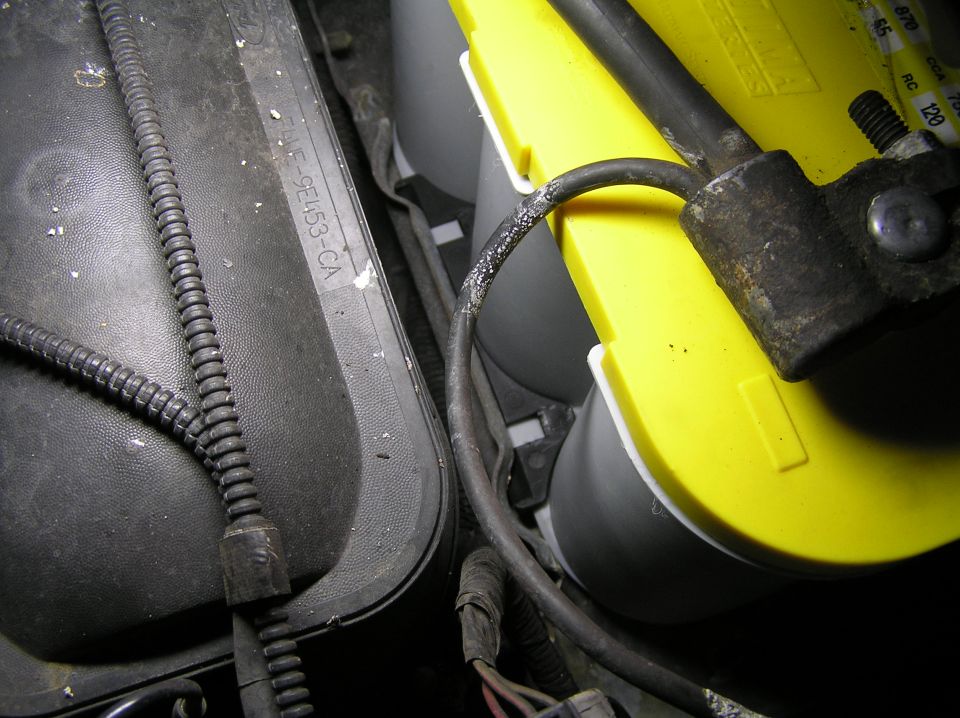 Yellow Optima battery with hold-down clamp next to a black engine bay component and wiring harness.