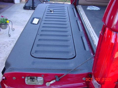 1989 Ford Bronco - Angled overhead shot of a red SUV's lowered tailgate, showing the ribbed gray plastic inner trim panel, latch striker and support cable, wi…