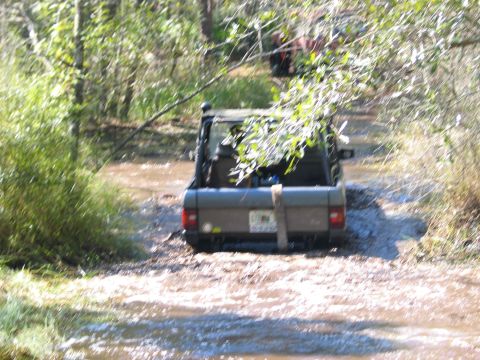 1992 Ford Bronco - A brown Ford Bronco navigates a muddy trail, with another vehicle visible in the background.