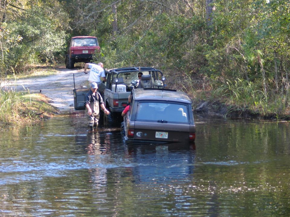 Group recovering vehicles from a flooded trail, one SUV deep in water, a red truck on dry ground