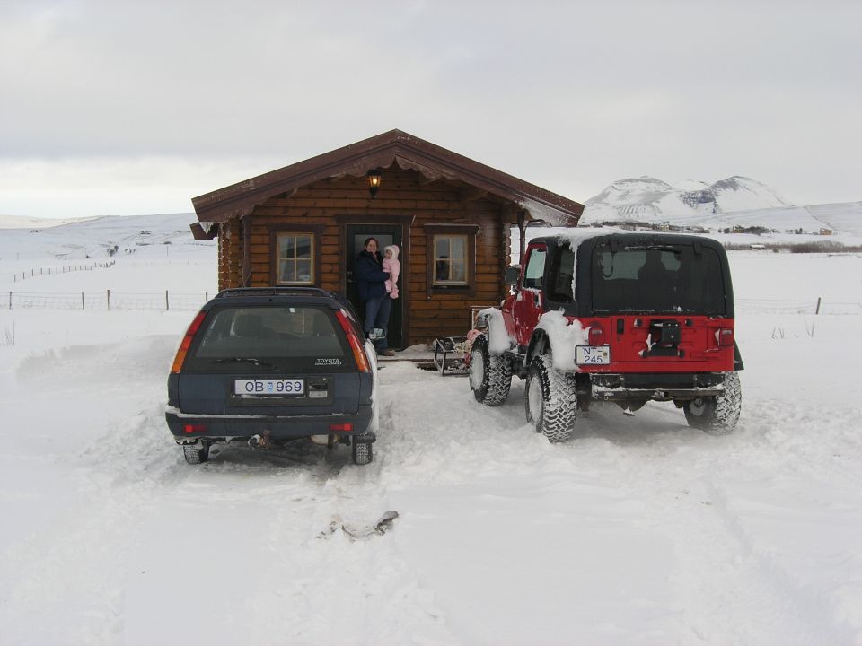 red Jeep Wrangler and blue station wagon parked in snow outside a wooden cabin. Two people and a child stand in the cabin doorway