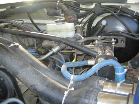 1990 Ford Bronco - Detailed engine bay showing the brake master cylinder reservoir, blue braided hoses, wiring looms and a wrapped exhaust manifold.