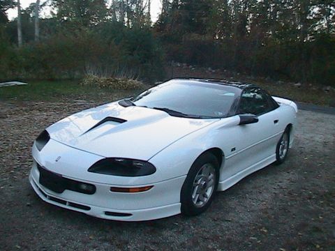 1996 Chevrolet Camaro - A white 1996 Chevrolet Camaro is parked on a gravel surface with trees and foliage behind it.