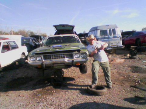 1979 Ford F-150 - A man stands next to a green 1974 Plymouth Barracuda coupe in a junkyard, with its hood open and front end elevated.