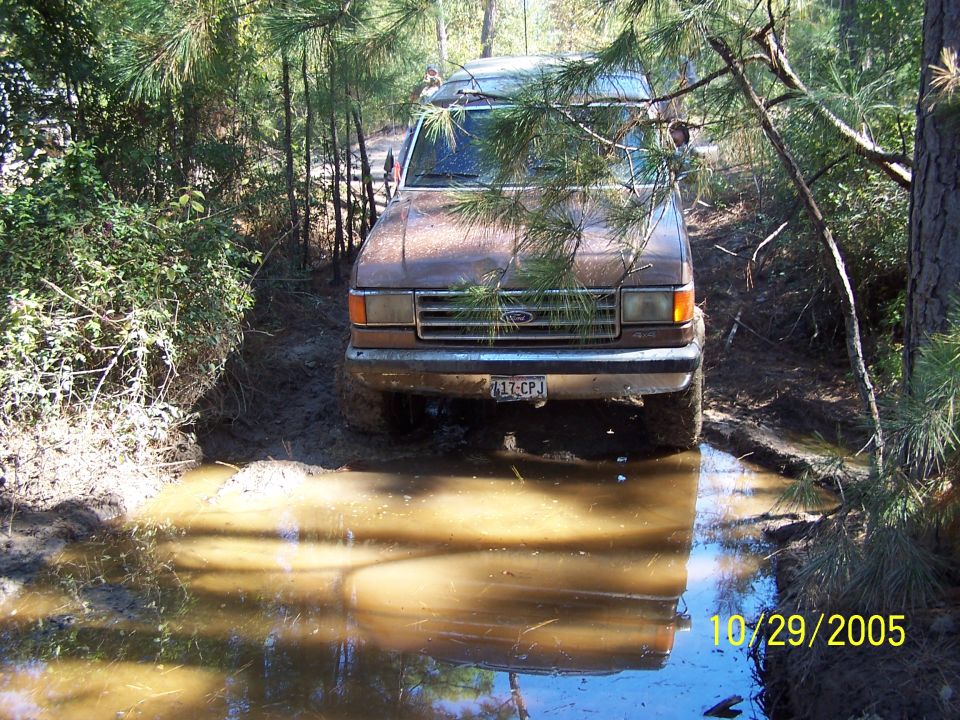 Brown 1987 Ford Bronco driving through a muddy trail, trees all around