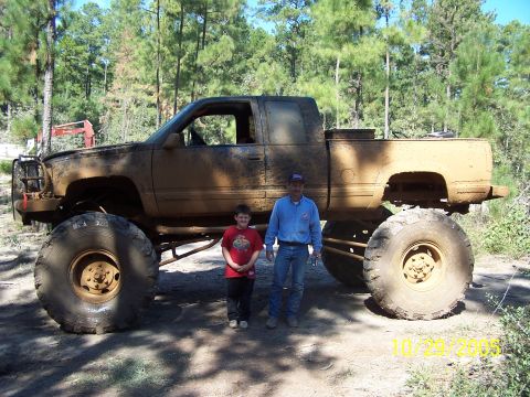 1987 Ford Bronco - A heavily modified pickup truck, covered in mud, stands tall with two people posing beside it in a wooded area.