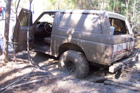1987 Ford Bronco - Mud-covered 1987 Ford Bronco stuck in a muddy wooded area with the driver's door open.
