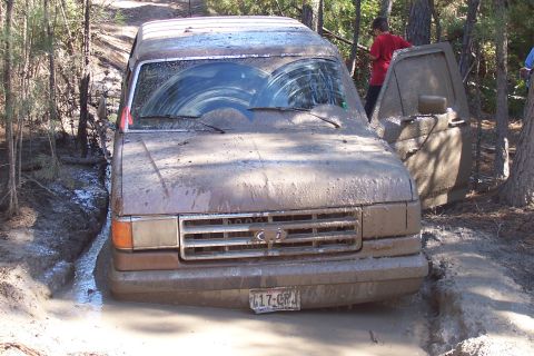 1987 Ford Bronco - A muddy 1987 Ford Bronco is stuck in a water-filled ditch with its driver's door ajar.