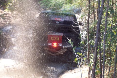 1987 Ford Bronco - A 1987 Ford Bronco navigates through mud and water, its rear end visible with a red spare tire cover.
