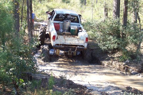 1987 Ford Bronco - A white pickup truck navigates a muddy off-road trail, its large tires deep in the mire.