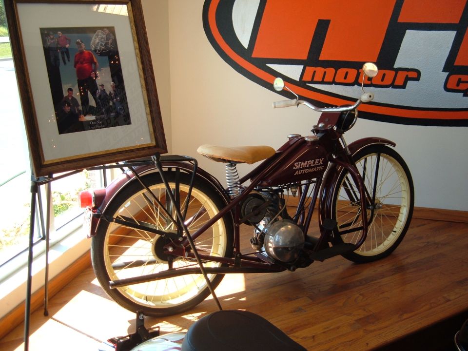 Maroon Simplex Automatic motorcycle with tan seat and cream wheels. Framed photo on stand nearby. Logo on wall