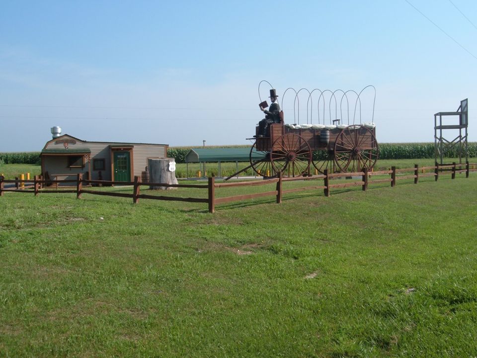 Old wagon with a figure in a top hat, next to a small building and a wooden fence
