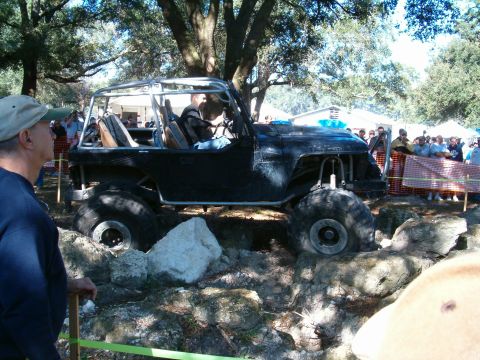 1991 Ford Bronco - A heavily modified Jeep with a roll cage and oversized tires is shown off-roading over rocks.