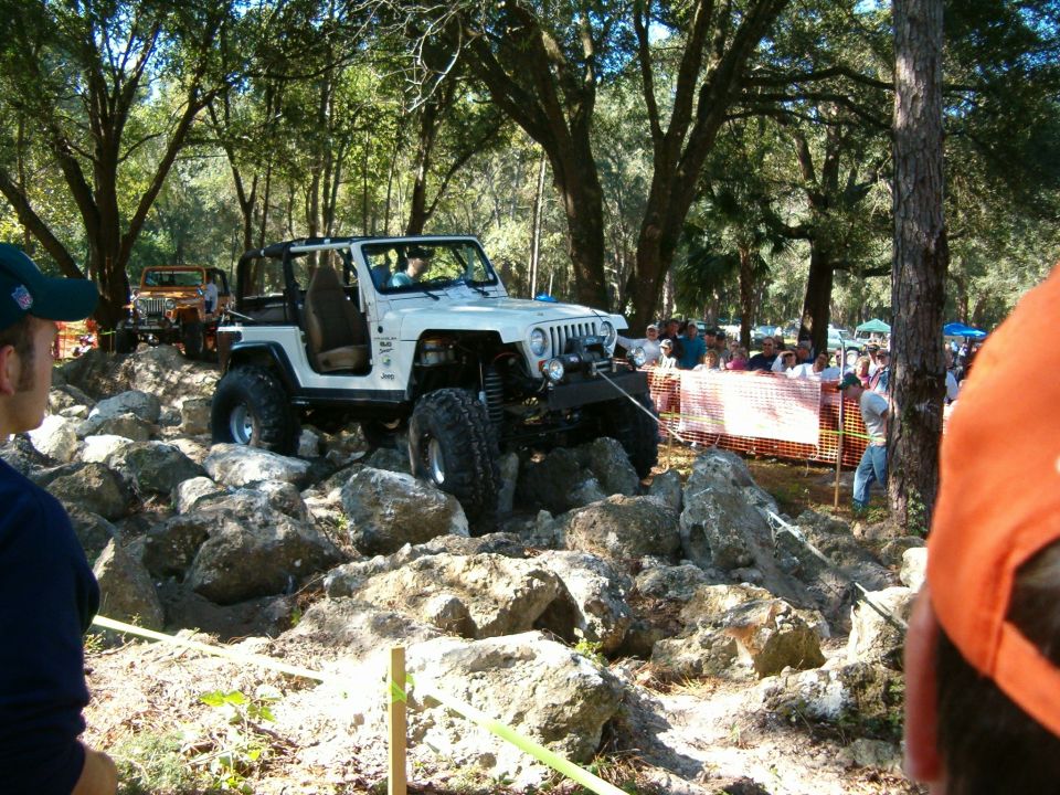 White Jeep Wrangler with large tires navigating a rock obstacle course, with spectators in the background