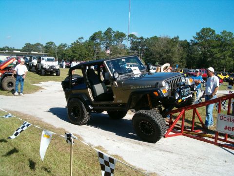 1991 Ford Bronco - A lifted Jeep Wrangler Rubicon is positioned on a red ramp at an outdoor event with other vehicles in the background.