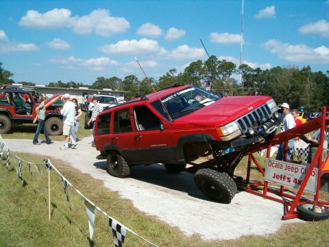 1991 Ford Bronco - A red Jeep Grand Cherokee is positioned on a ramp at an off-road event, with other vehicles and spectators in the background.