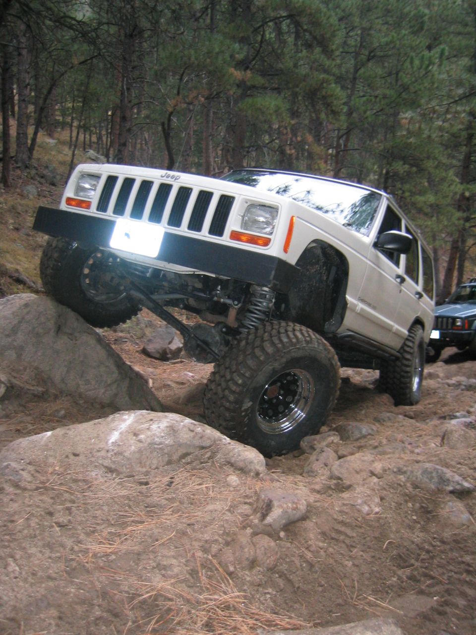 White Jeep Cherokee climbing over rocks on a wooded trail. Another Jeep is visible in the background