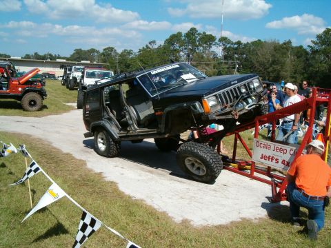 1991 Ford Bronco - A modified Jeep Cherokee XJ is positioned on a ramp at an event, with other vehicles and spectators in the background.