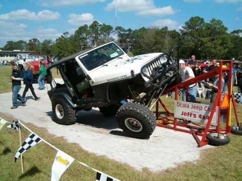 1991 Ford Bronco - Jeep Wrangler tackles an obstacle course at an event with other vehicles and spectators present.